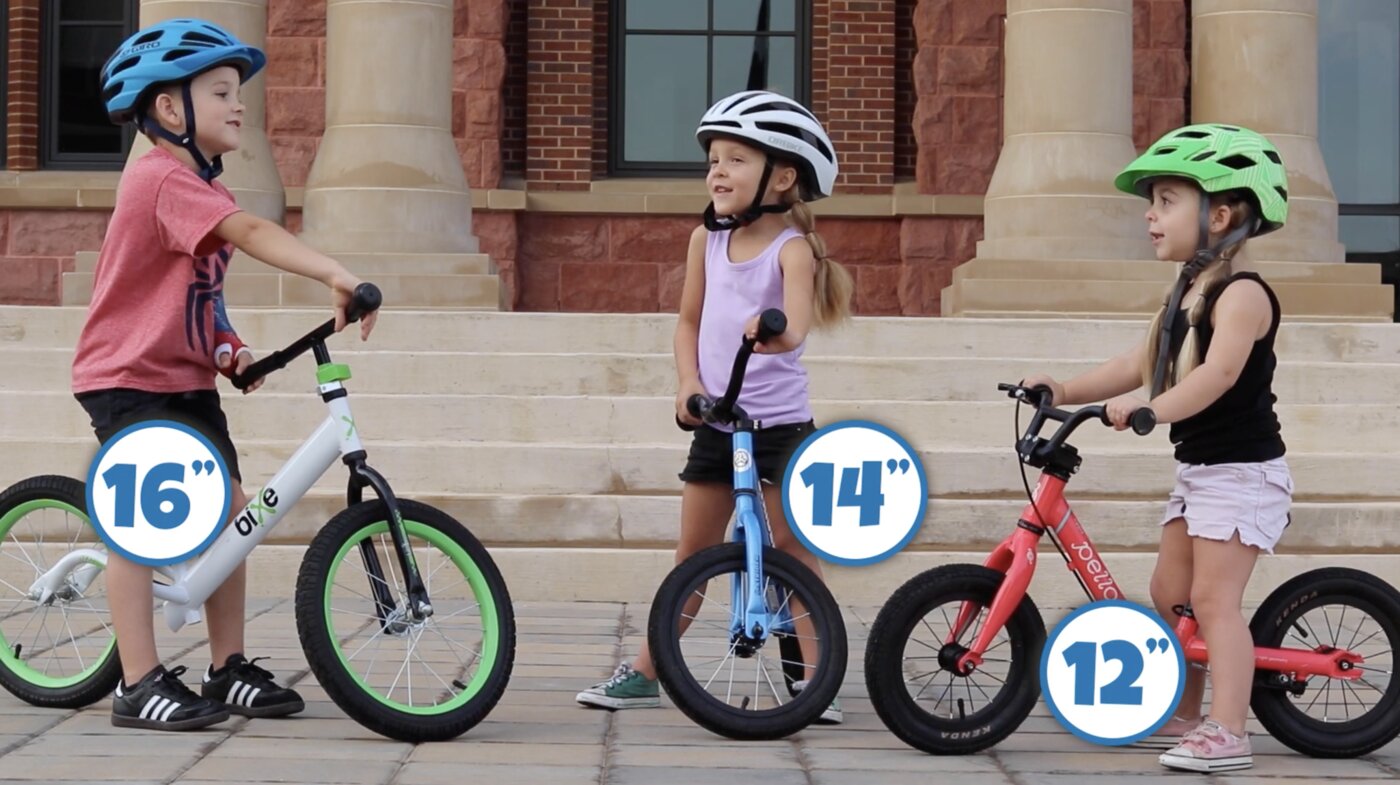 Drie kinderen op loopfietsen met wielmaten van 16, 14 en 12 inch naast elkaar, ter vergelijking van de formaten