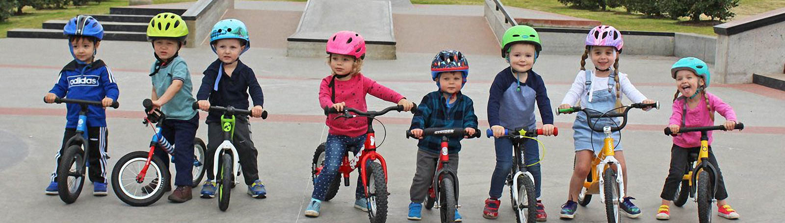 Acht kinderen met uiteenlopende loopfietsen en gekleurde helmen op een skatepark
