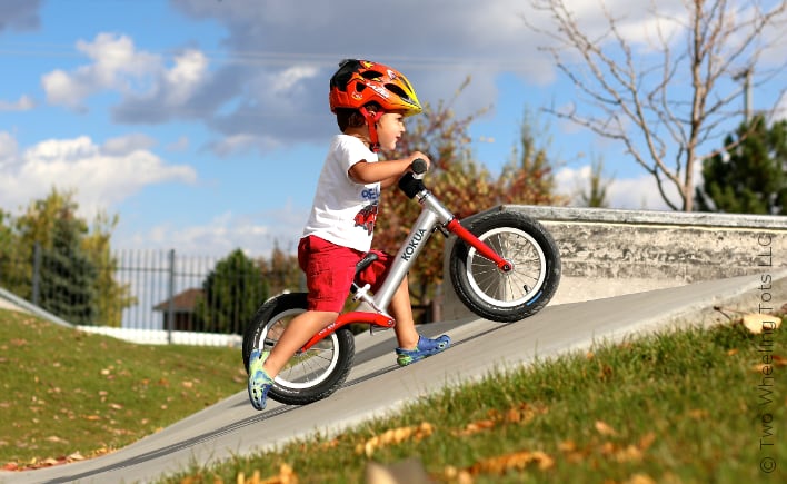 Jongen met helm rijdt op een rode loopfiets een helling af op een skatepark