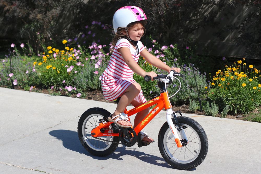 Meisje in gestreepte jurk fietst zelfstandig op een oranje kinderfiets zonder zijwieltjes langs een bloemenperk