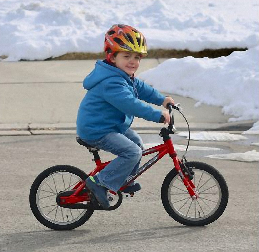 Jongen in blauwe hoodie rijdt op een rode kinderfiets zonder zijwieltjes door een besneeuwde straat