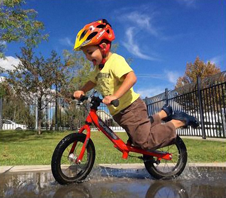 Jongen trekt lachend zijn voeten op tijdens het rijden op een rode loopfiets door een plas water